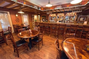 A bar area with wooden tables and chairs, a bar counter with bottles, and a ship's wheel on the wall.