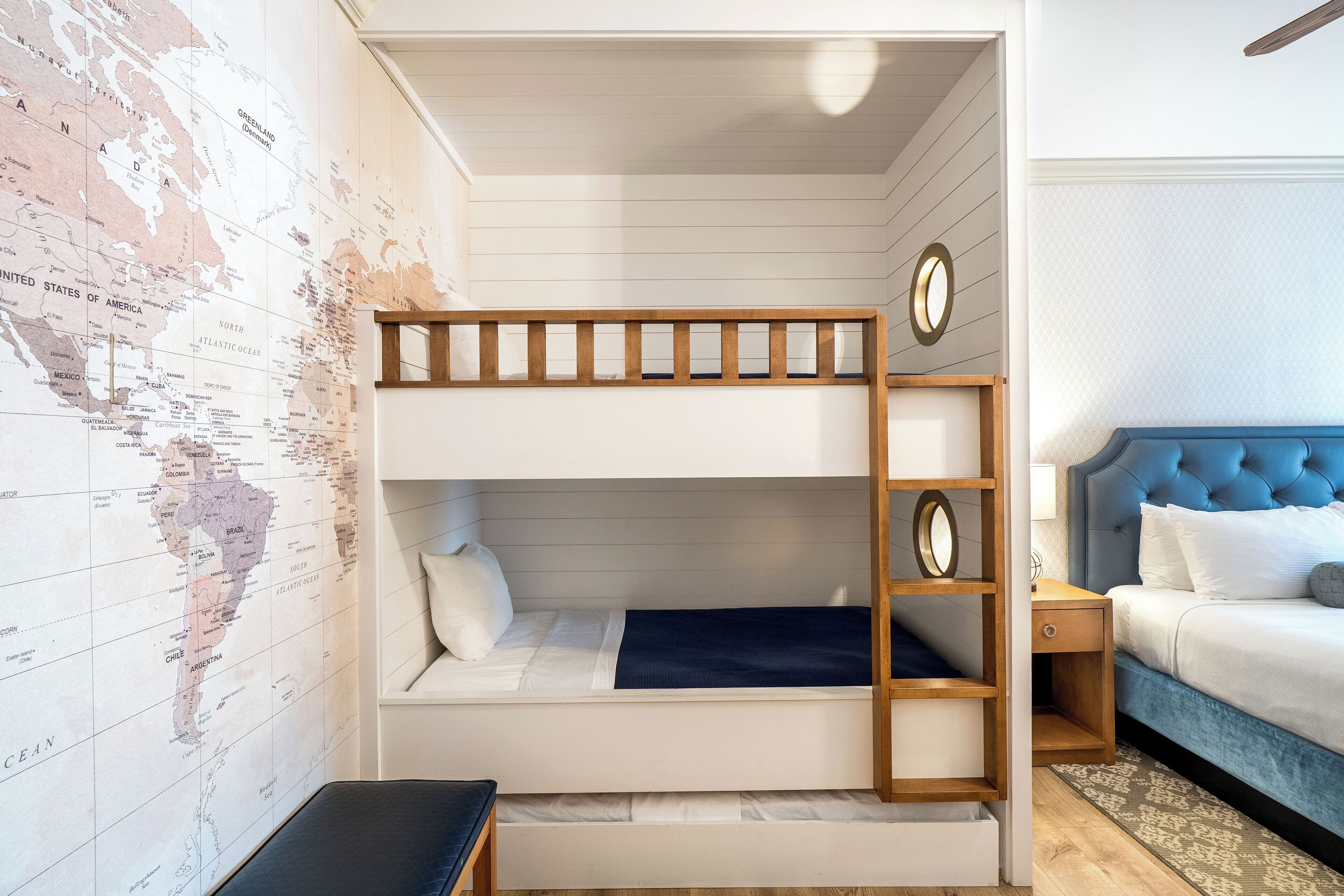 Uma cama beliche com escada de madeira, uma pequena escrivaninha e um mapa-múndi na parede.