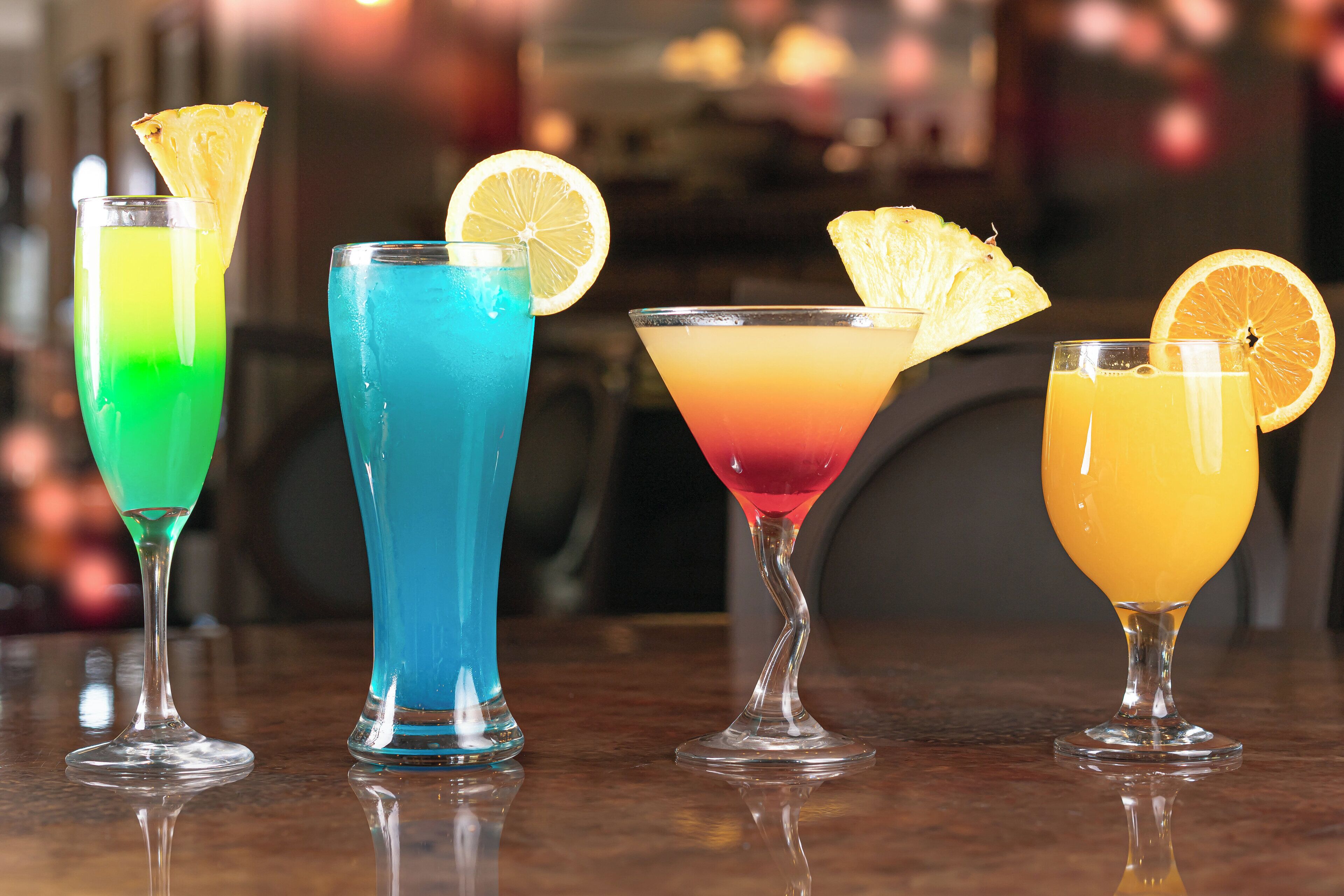 Four colorful cocktails with garnishes on a reflective surface.