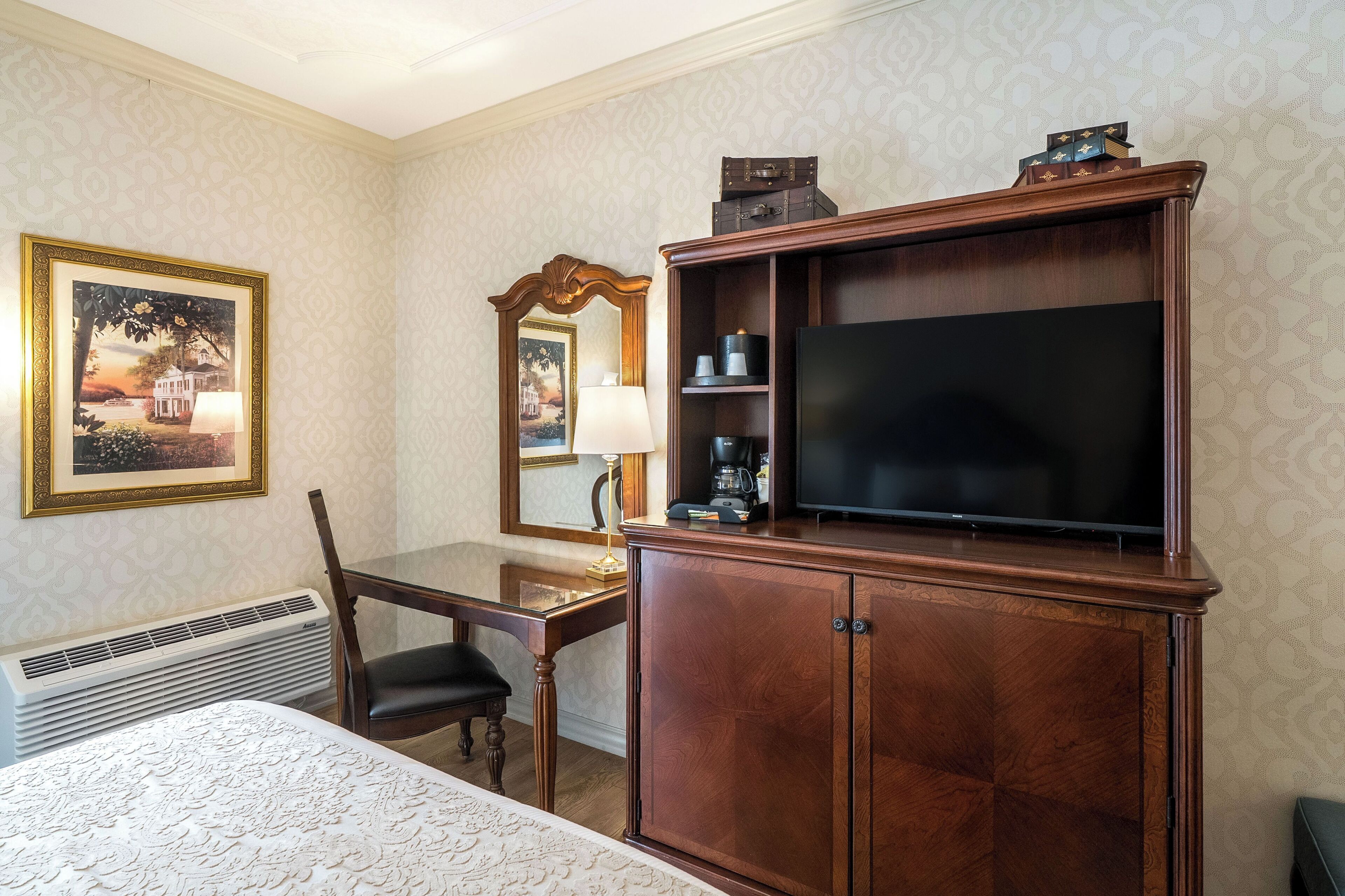 A hotel room with a bed, a wooden cabinet with a television, a desk with a chair, a mirror, and a framed picture on the wall.