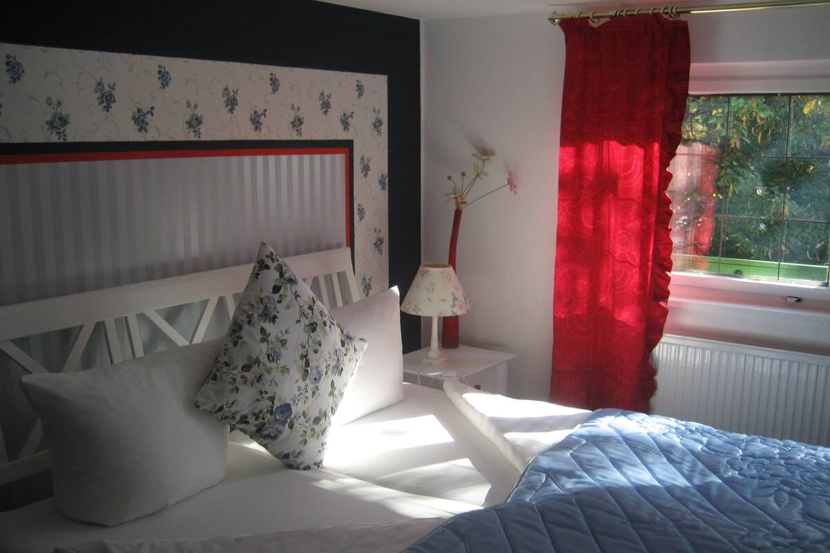 A bedroom with a bed, white headboard, floral pillows, a bedside table with a lamp, and a window with red curtains.