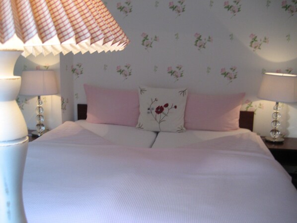 Una cama bien hecha con ropa de cama rosa y blanca, dos lámparas de noche y papel tapiz floral.