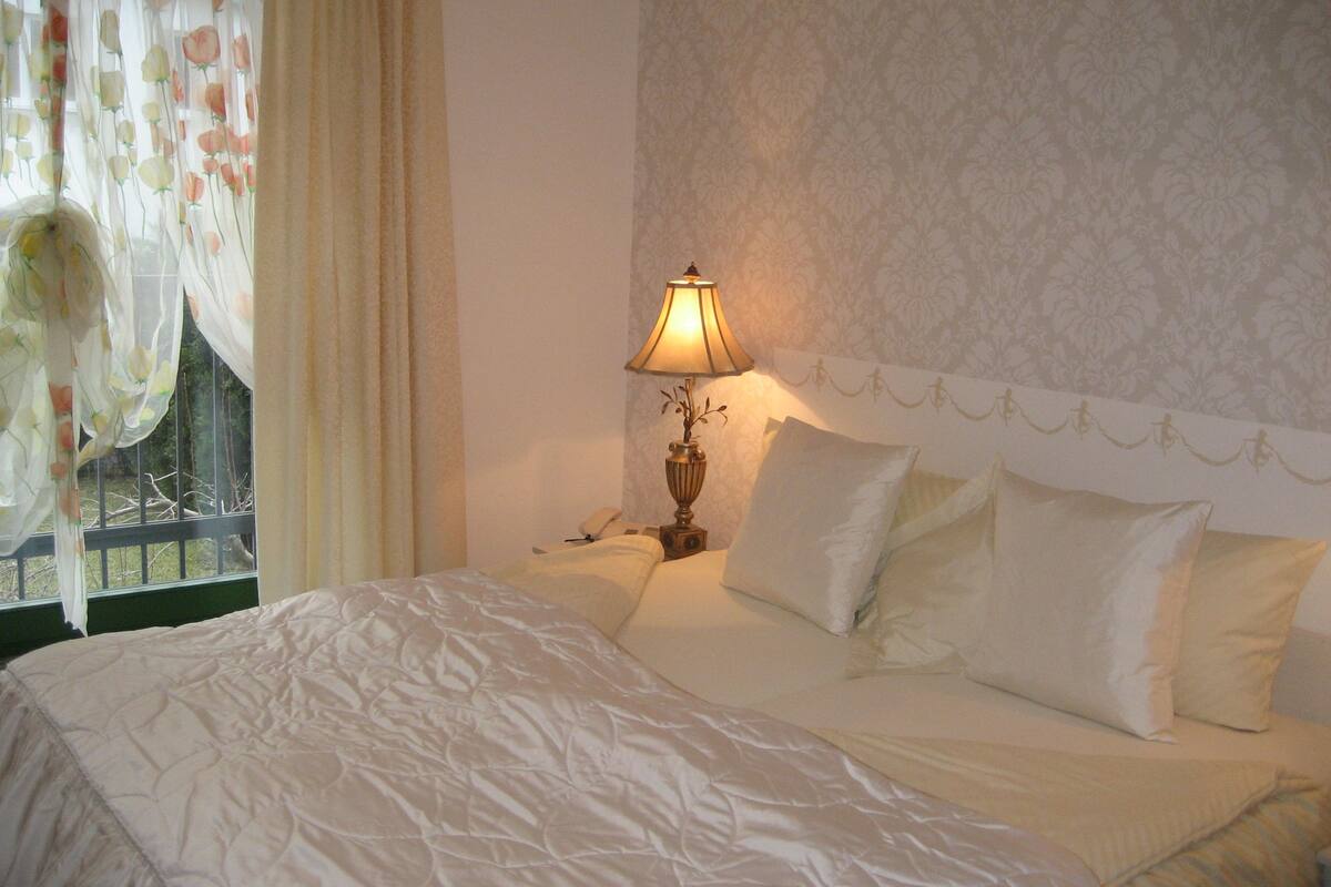 A neatly made bed with white linens, a bedside lamp, and a floral curtain.