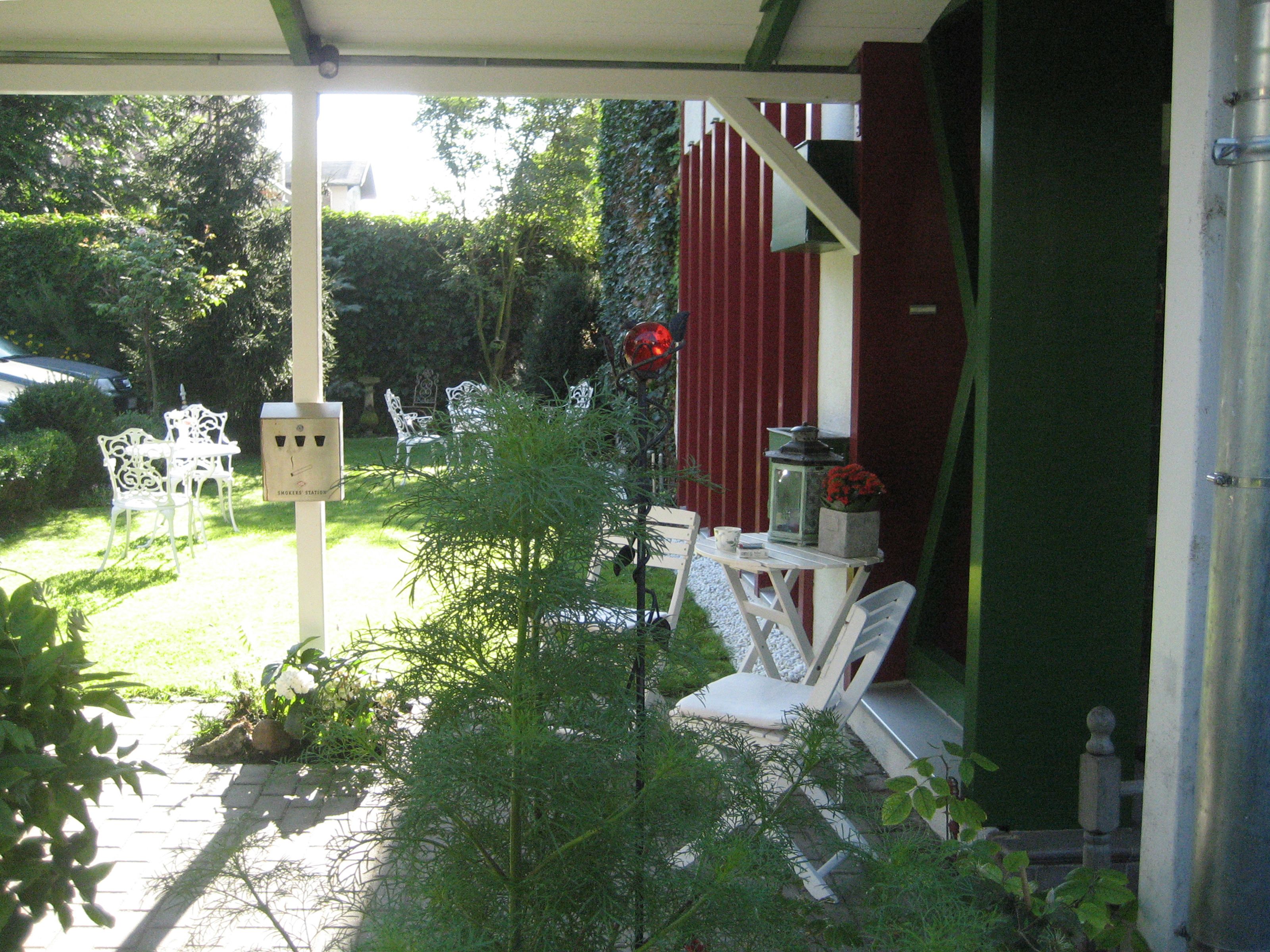 A covered outdoor seating area with white chairs and a table, surrounded by greenery and a red building in the background.