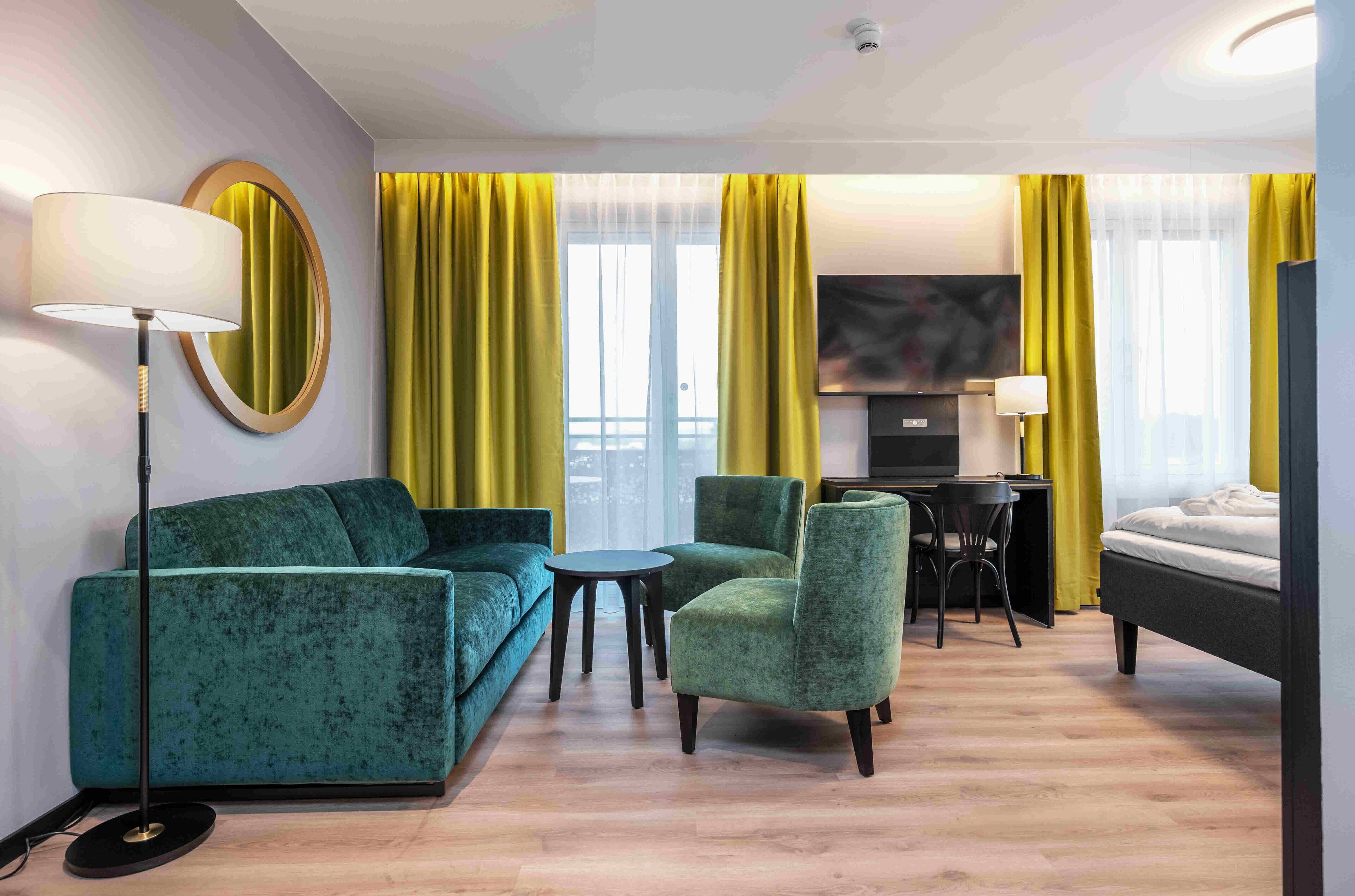 A hotel room with a green sofa, a small round table, a green armchair, a bed, a desk with a chair, and a TV.