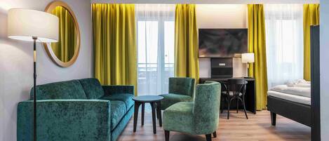 A hotel room with a green sofa, a small round table, a green armchair, a bed, a desk with a chair, and a TV.