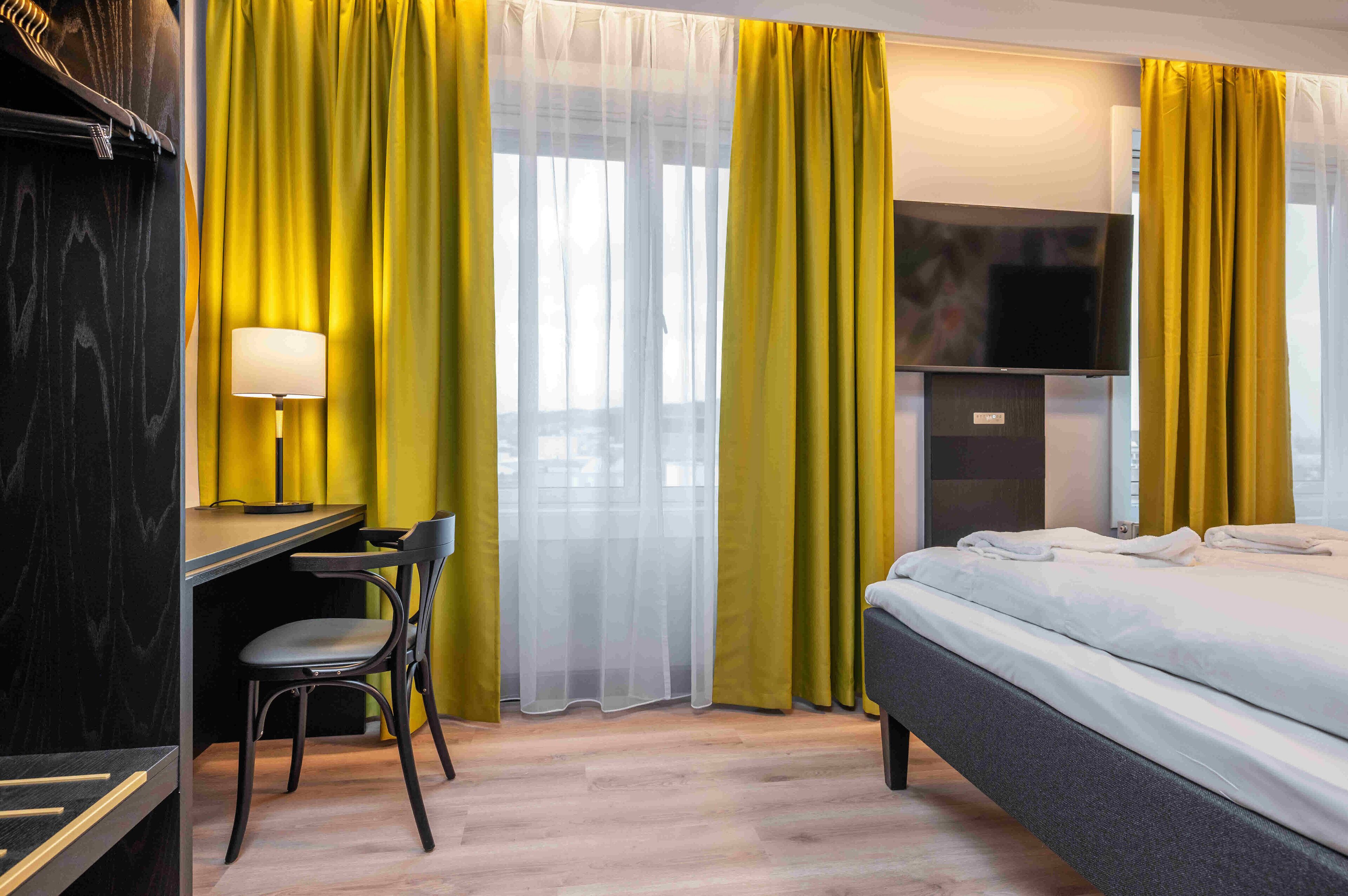 A hotel room with a bed, a desk with a chair, a lamp, and yellow curtains.