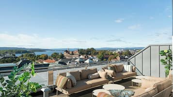 Una terraza con mobiliario exterior, vista a un paisaje urbano y un cuerpo de agua a lo lejos.