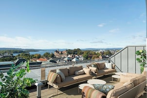 Una terraza con mobiliario exterior, vista a un paisaje urbano y un cuerpo de agua a lo lejos.