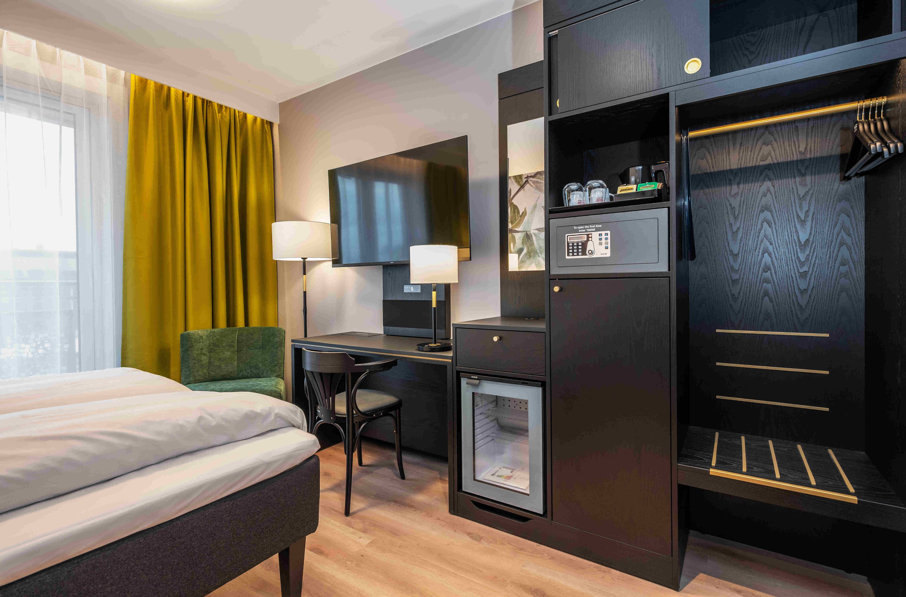 A hotel room with a bed, desk, chair, television, lamp, minibar, and wardrobe.