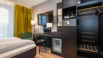 A hotel room with a bed, desk, chair, television, lamp, minibar, and wardrobe.