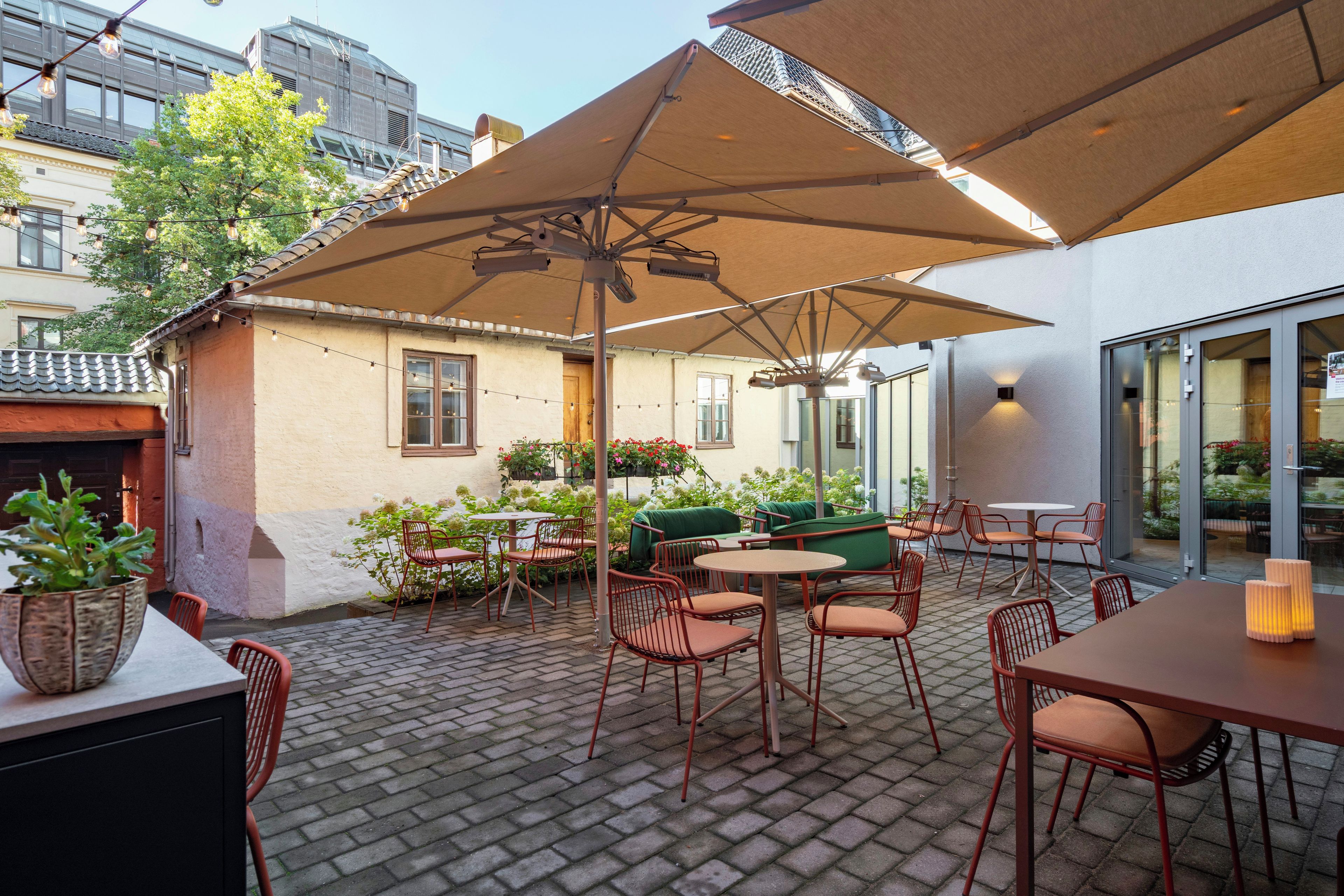 An outdoor patio area with brick flooring, red metal chairs, and tables, featuring a large umbrella and potted plants.