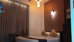A modern hotel room with a large bed, a small table, and a chair. There is a curtain, a wall-mounted light, and a ceiling light. - Silicrest Hotel (Bengaluru)