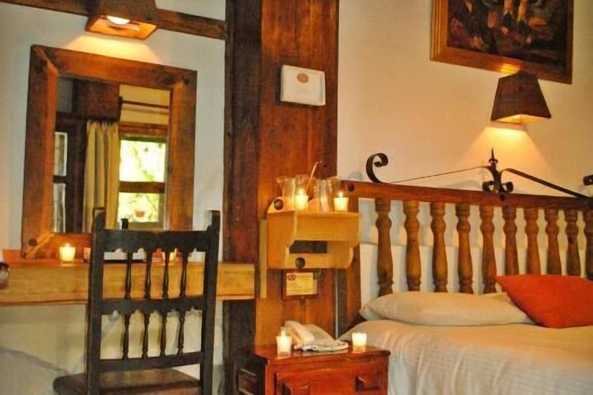 A cozy room with a wooden bed, a chair, a small table with candles, and a painting on the wall.