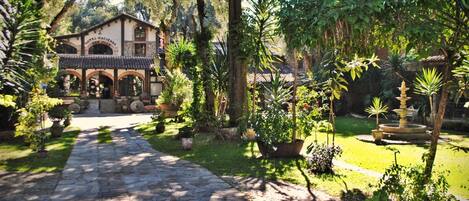 Un hotel de estilo arquitectónico tradicional, un sendero de piedra que lleva a la entrada y un jardín cuidado con diversas plantas y una fuente.