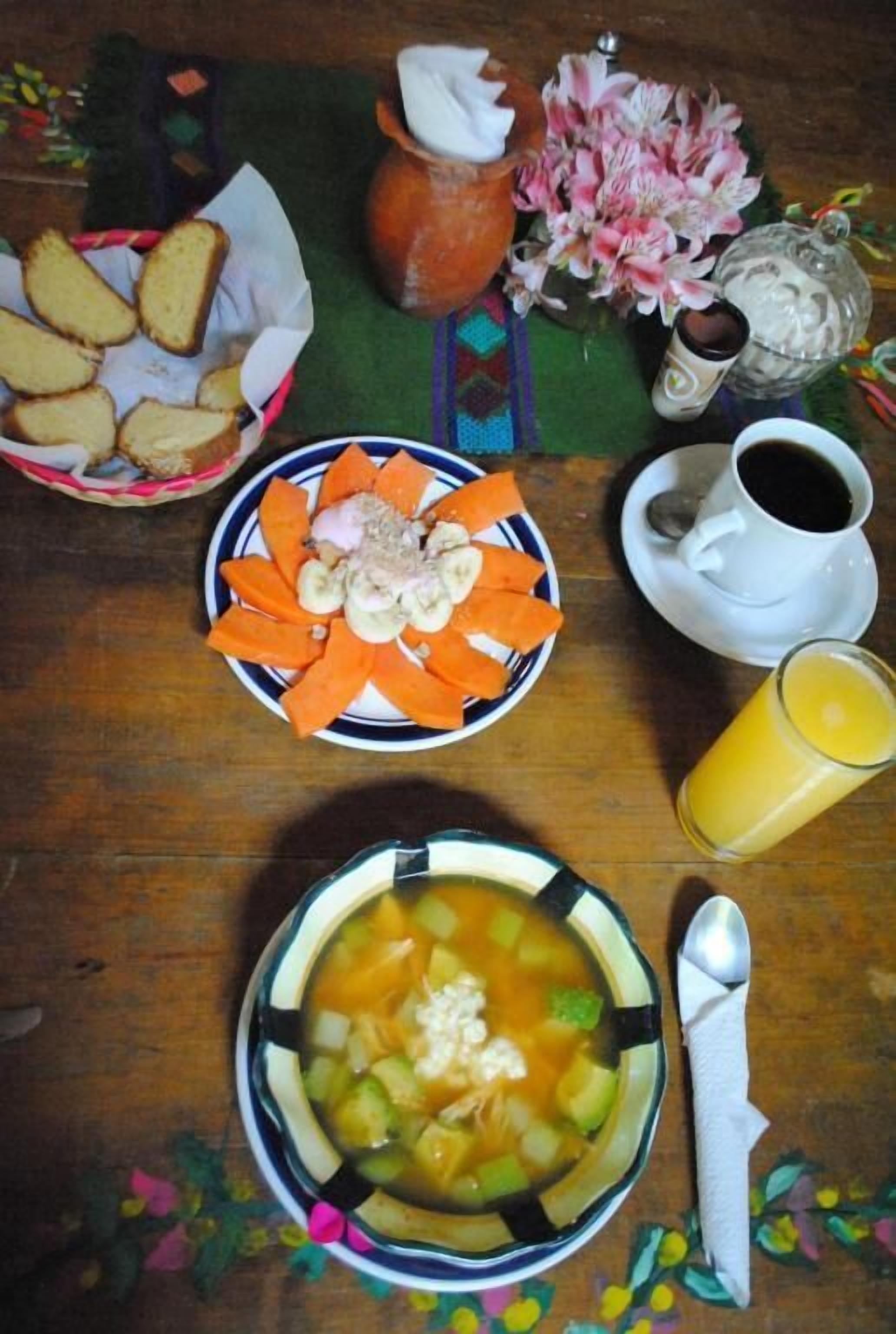 Uma mesa com uma travessa de frutas fatiadas, uma tigela de sopa, uma xícara de café, um copo de suco e uma cesta de pão.