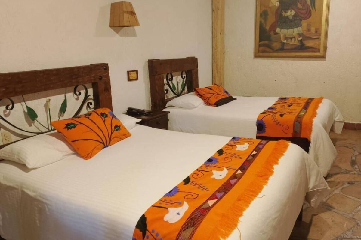 Two beds with matching headboards and orange decorative pillows, a wall-mounted lamp, and a painting above the beds.