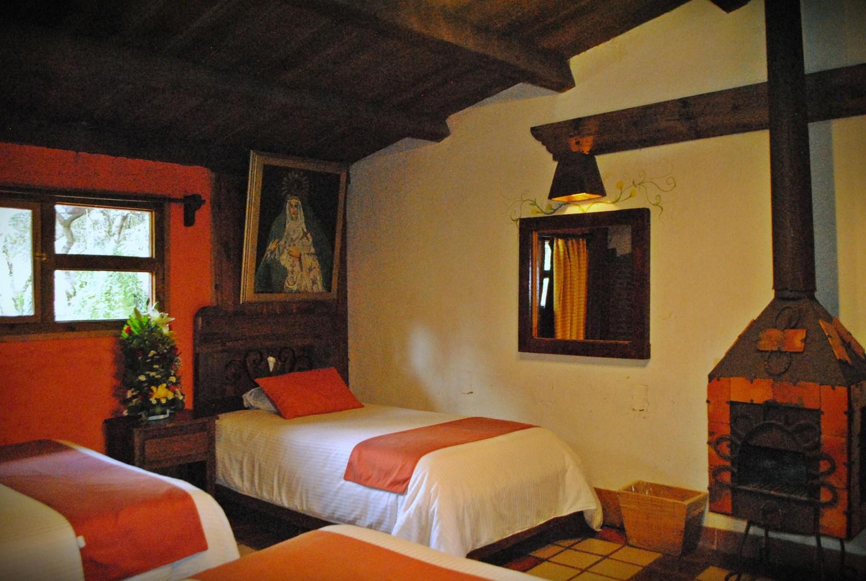 A room with two beds, a wooden ceiling, a fireplace, and a window with a view of trees.