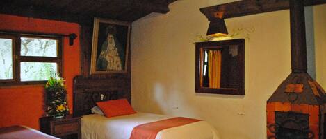 A room with two beds, a wooden ceiling, a fireplace, and a window with a view of trees.