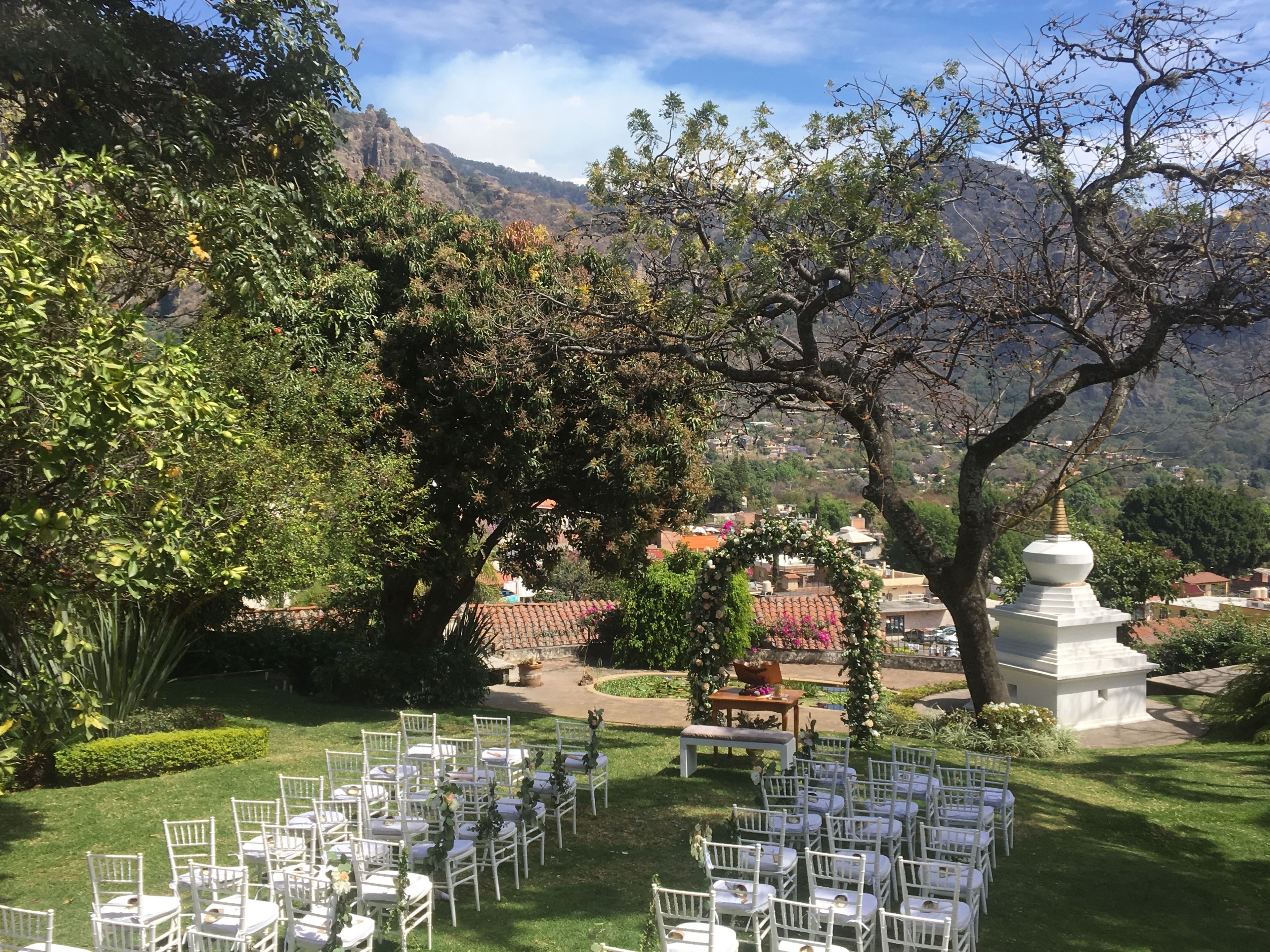 An outdoor wedding venue with rows of white chairs and tables, surrounded by lush greenery and a mountainous backdrop.