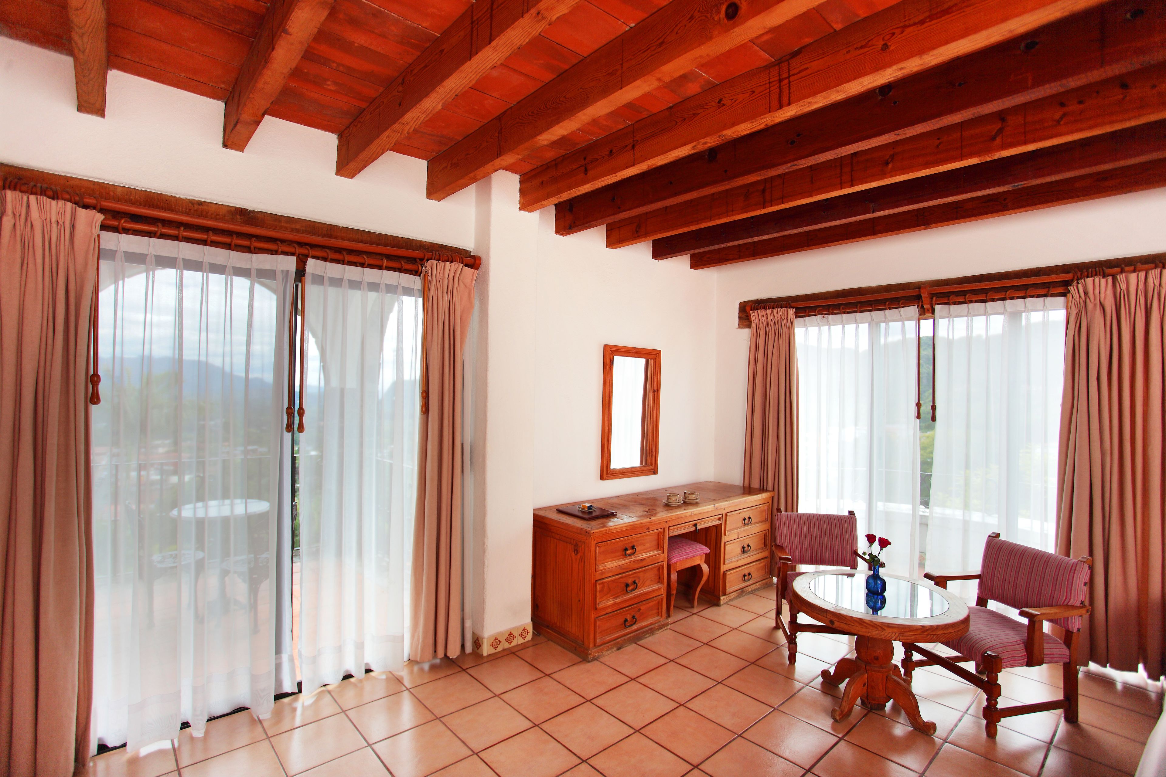 A room with a wooden ceiling, a desk with drawers, a chair, a round table, and two windows with curtains.