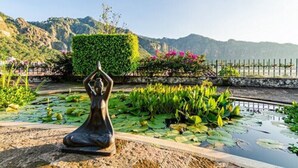 Una estatua de una persona en posición meditativa en un jardín con nenúfares y montañas al fondo.