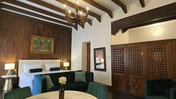 Habitación de hotel con cama, sofá, mesa de comedor con un jarrón de flores, candelabro, vigas de madera, un cuadro y una puerta de madera con celosías.