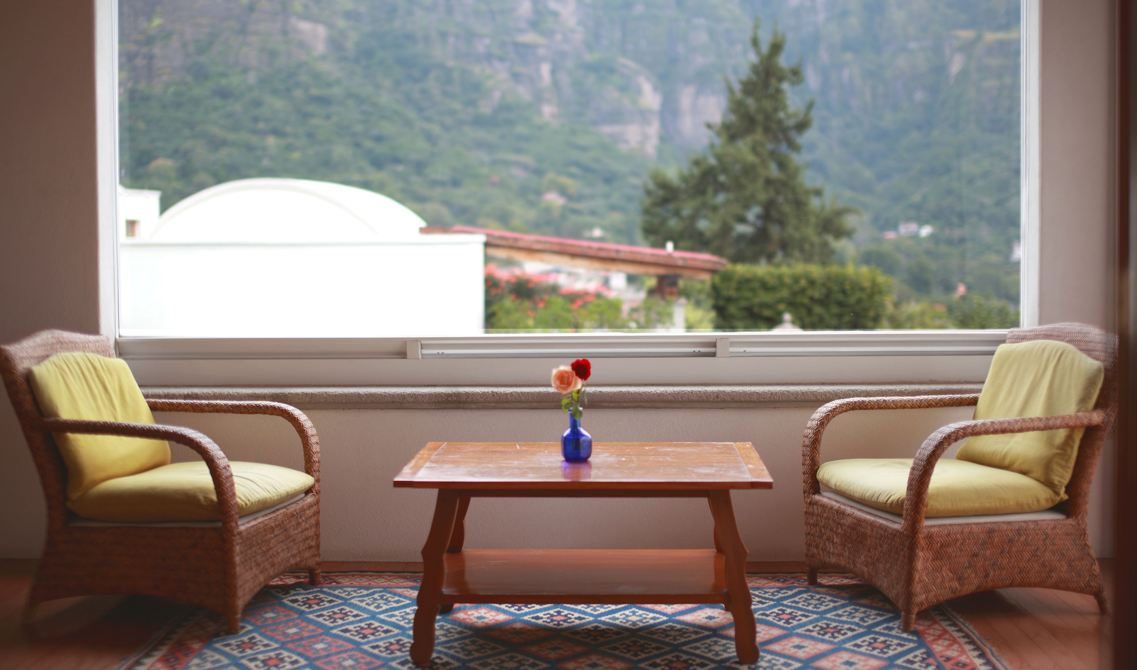A room with two wicker chairs, a small table, and a window with a view of mountains and buildings.