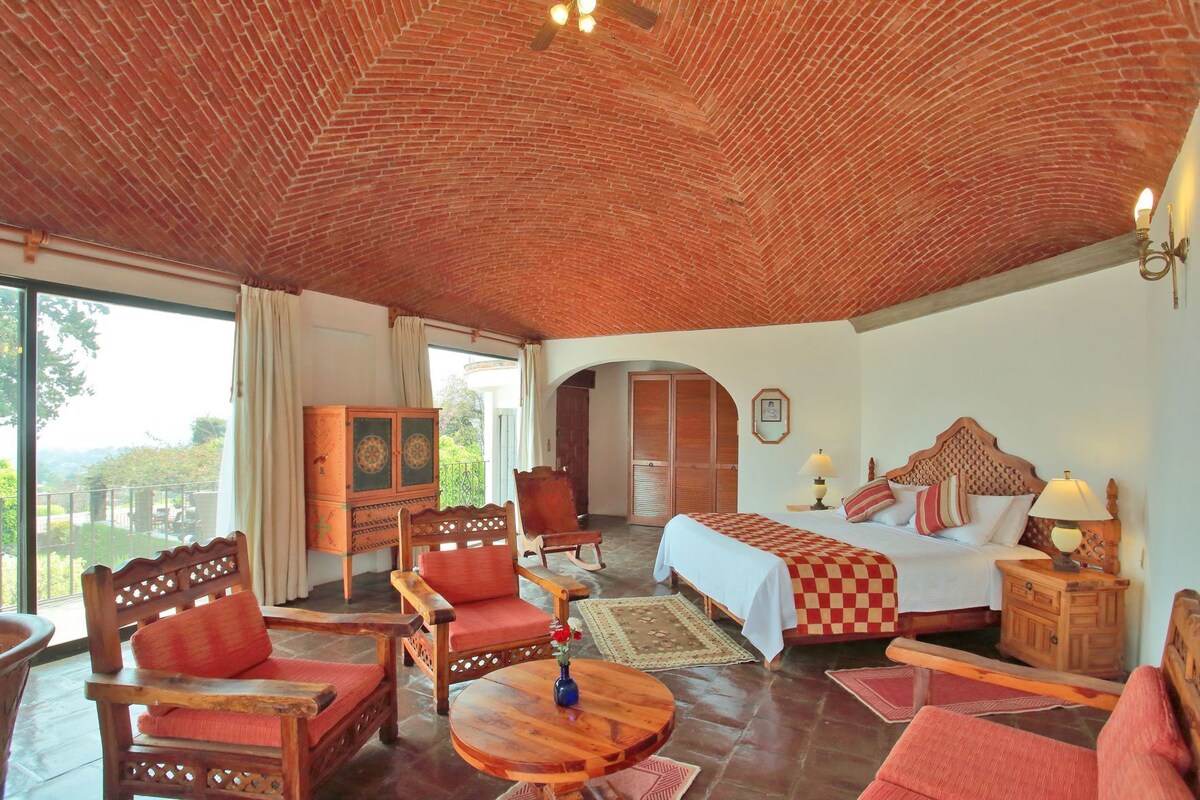 A room with a checkered bedspread, wooden furniture, a view through a large window, and a brick ceiling.