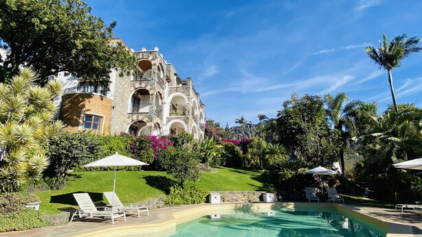 Un edificio grande de piedra con varios balcones y una piscina rodeada de abundante vegetación y palmeras.