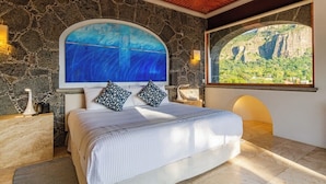 Un dormitorio con pared de piedra, una cama grande con almohadas estampadas, una mesita de noche, una lámpara y vistas a un paisaje montañoso a través de una ventana.