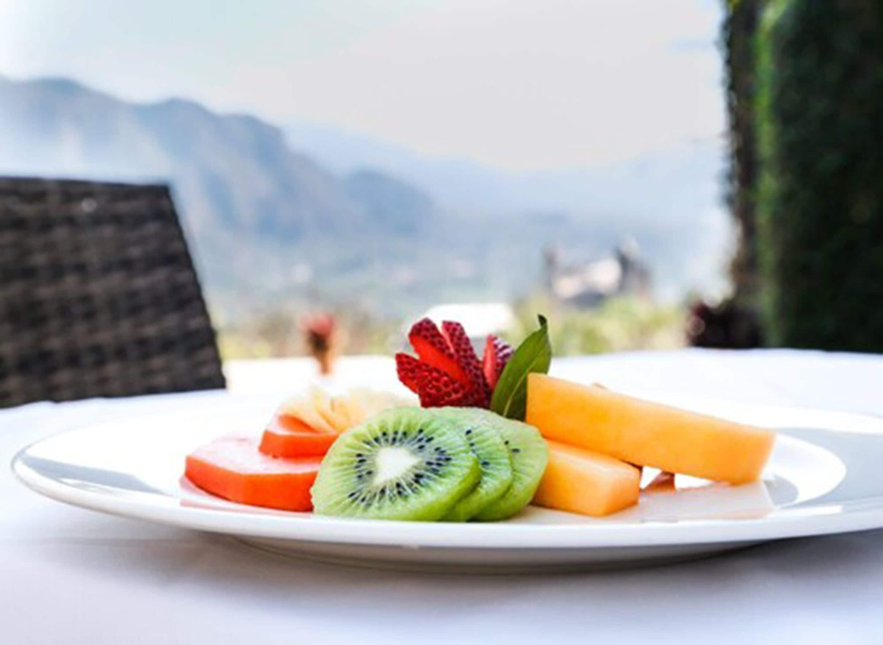 Prato com frutas fatiadas, incluindo kiwi, morangos e melancia, sobre uma mesa com vista para montanhas ao fundo.