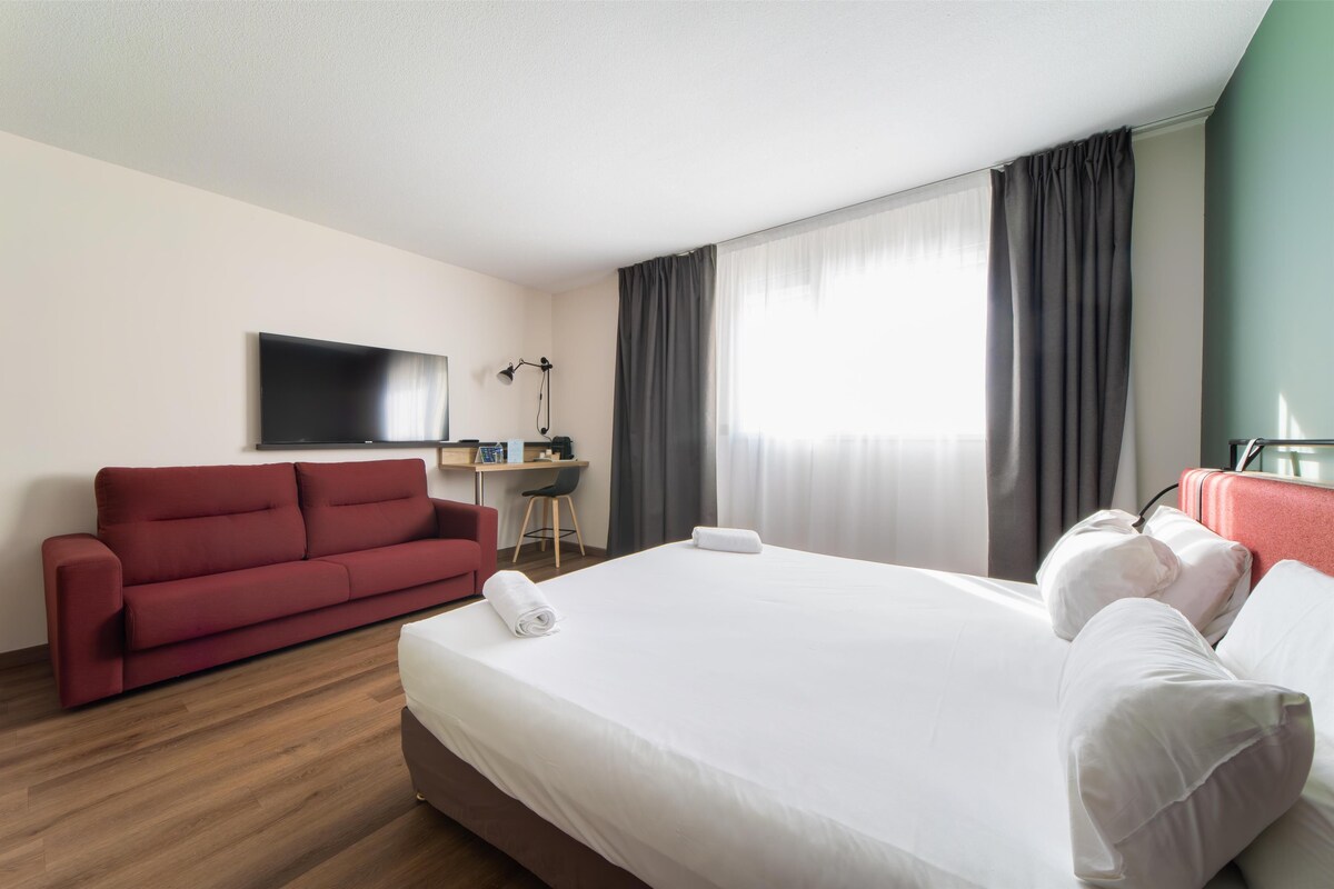 A hotel room with a large bed, a red sofa, a desk with a chair, and a TV mounted on the wall.
