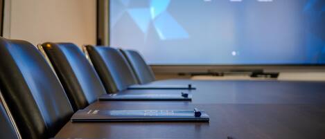 A conference room with a long table, black leather chairs, and a digital screen displaying the time.