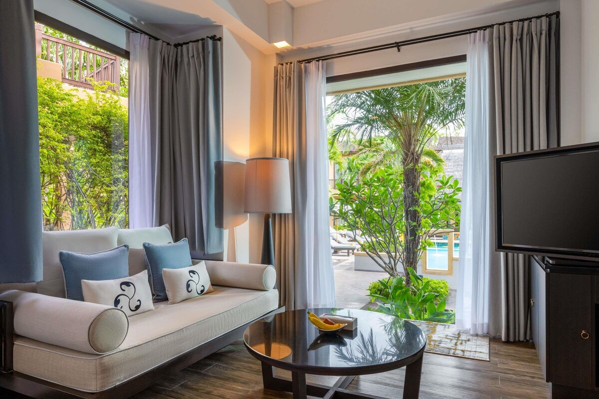 A modern hotel room with a sofa, a coffee table, a TV, and a view of a pool and greenery.