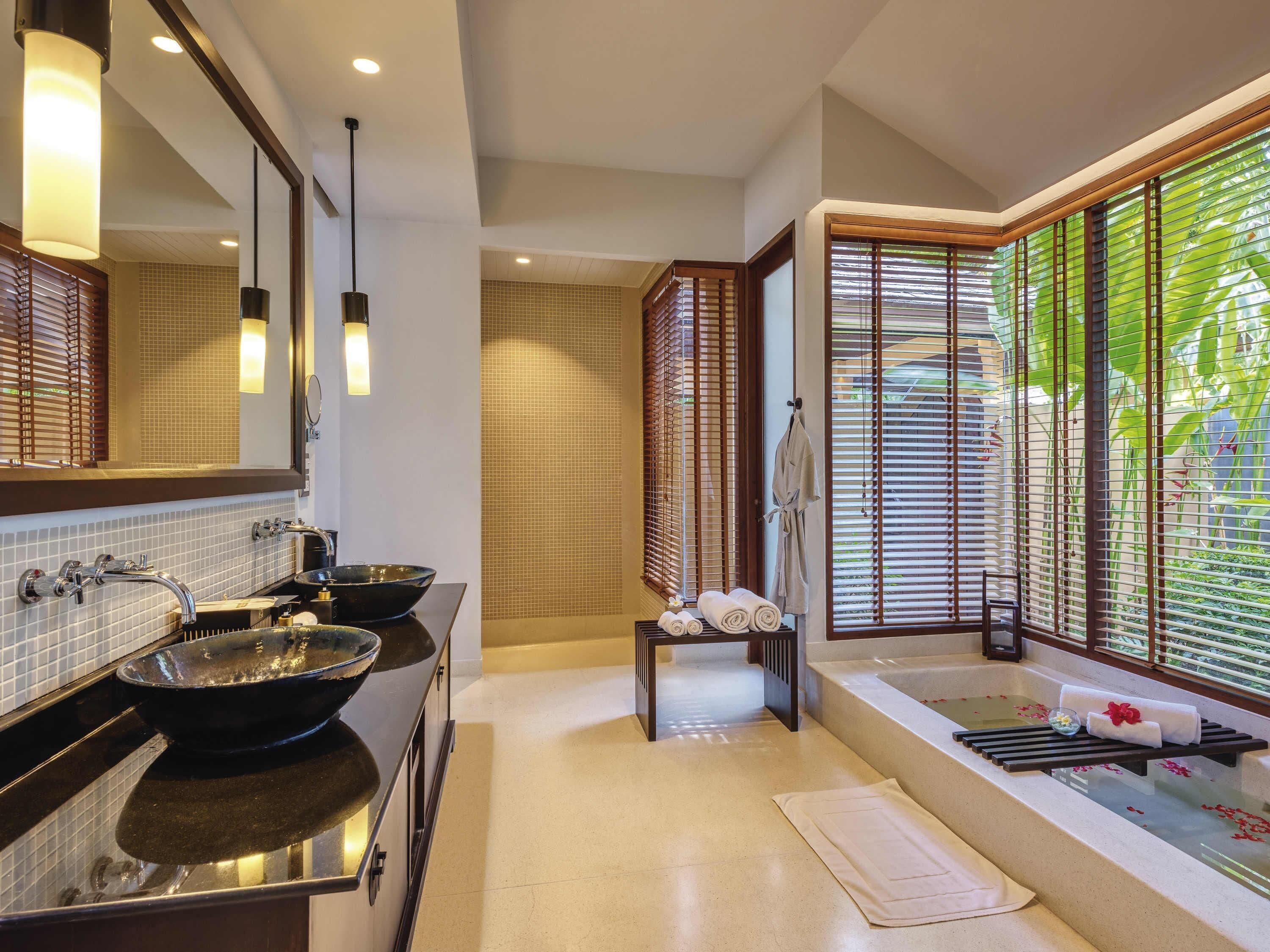 A modern bathroom with a large mirror, two sinks with black bowls, a bathtub, and a window with blinds.
