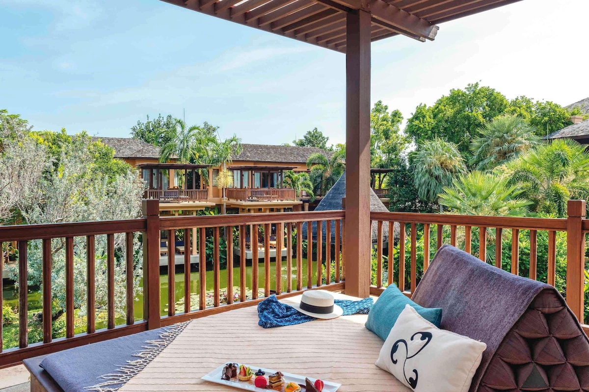 Un balcón con cubierta de madera, una mesa de comedor con una fuente de postres y vistas a un edificio de resort rodeado de vegetación.