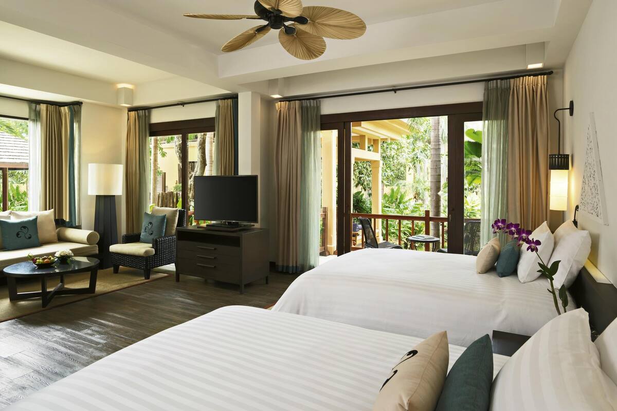 A hotel room with two beds, a desk, a television, and a balcony with a view of greenery.
