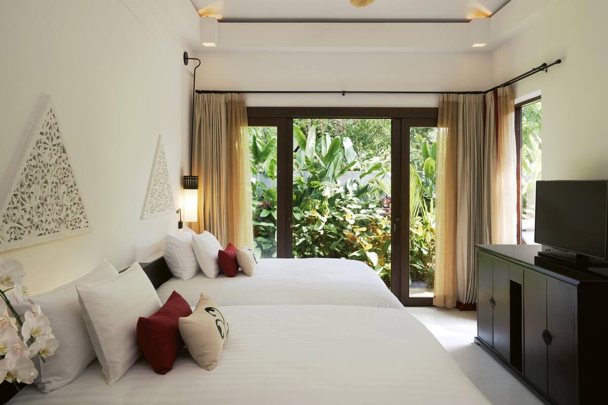 A modern hotel room with two beds, a television, a ceiling fan, and a view of lush greenery outside.