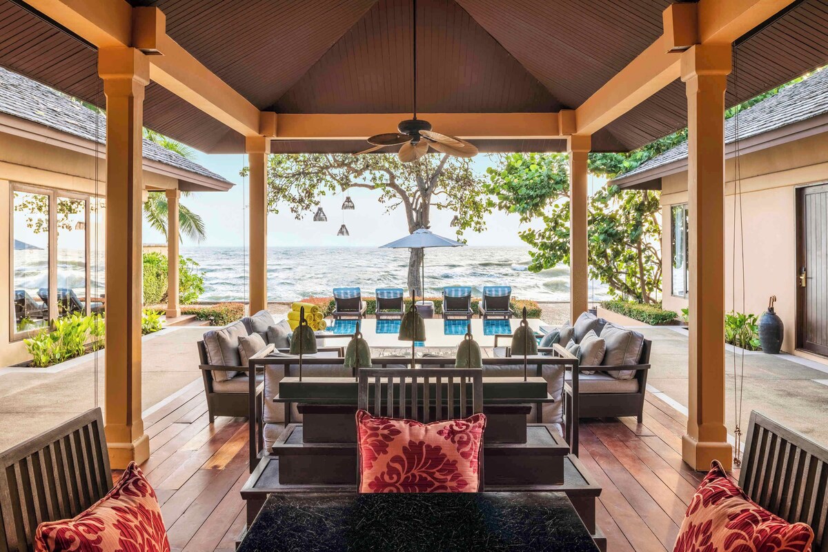 A covered outdoor seating area with a pool, lounge chairs, and a view of the ocean.