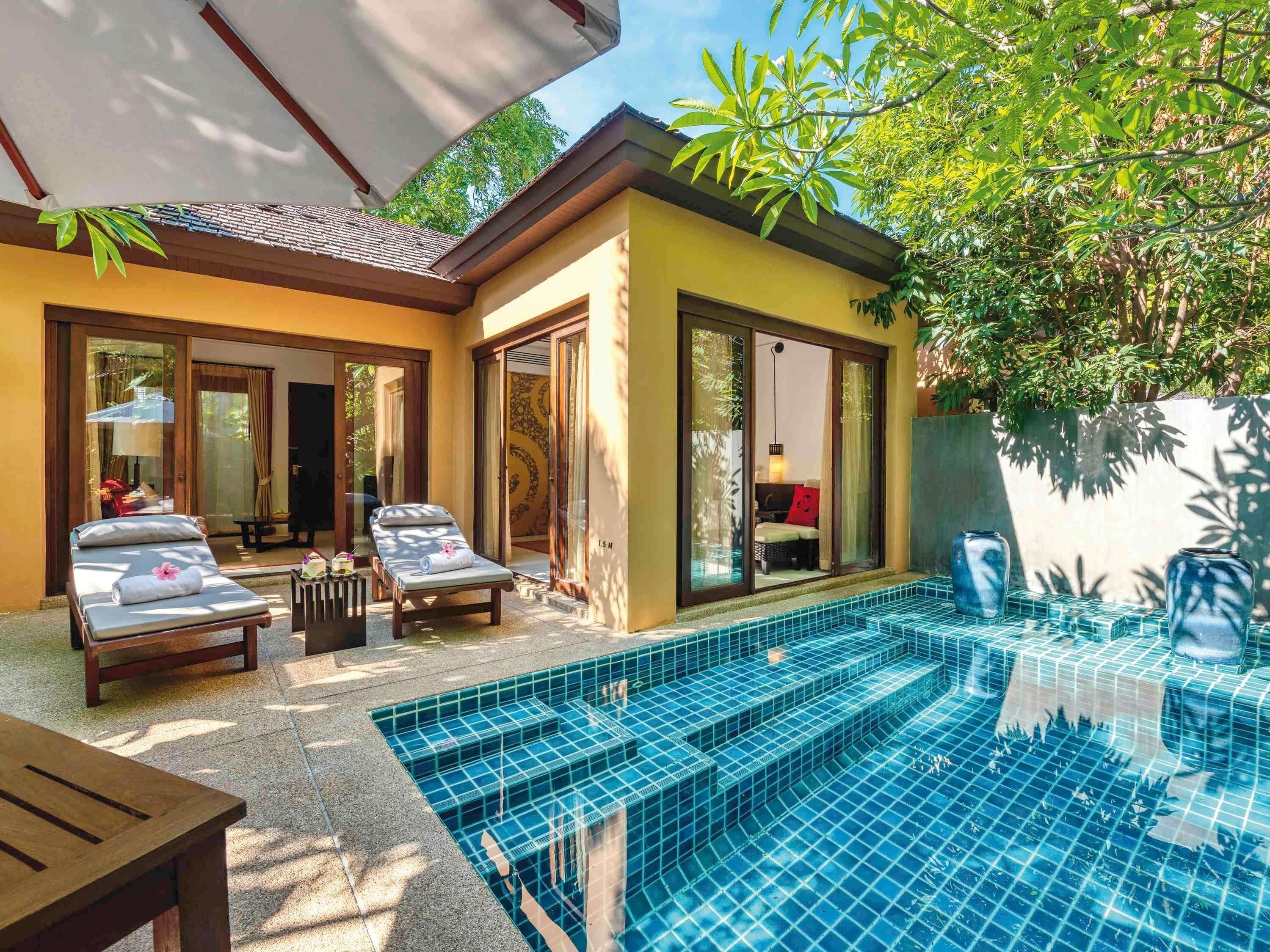 A poolside area with two lounge chairs, a wooden table, and a glass-enclosed room with a bed.