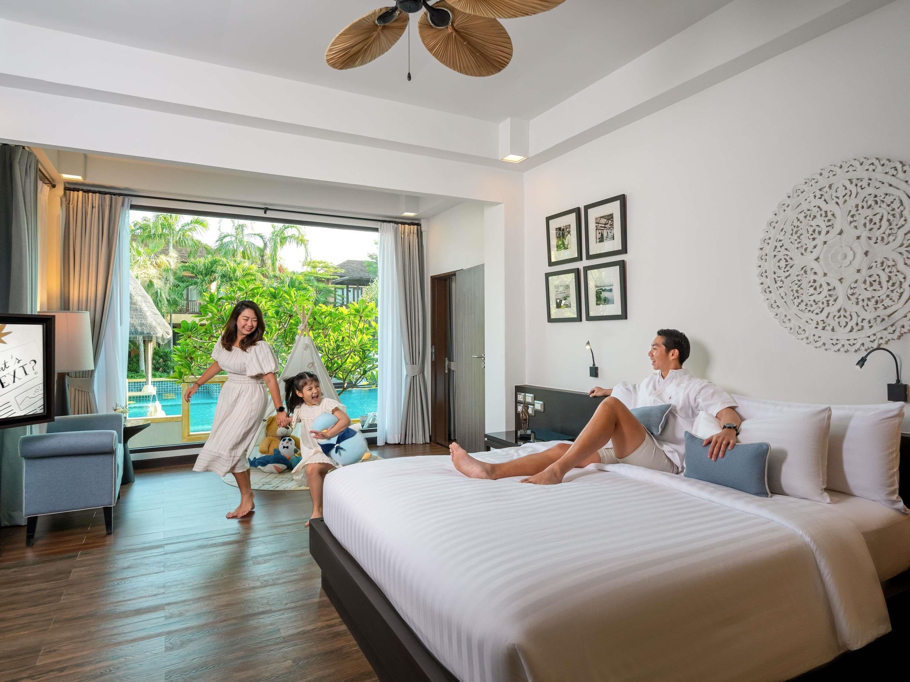 A hotel room with a bed, a desk, a chair, a window with a view of greenery, and a ceiling fan.