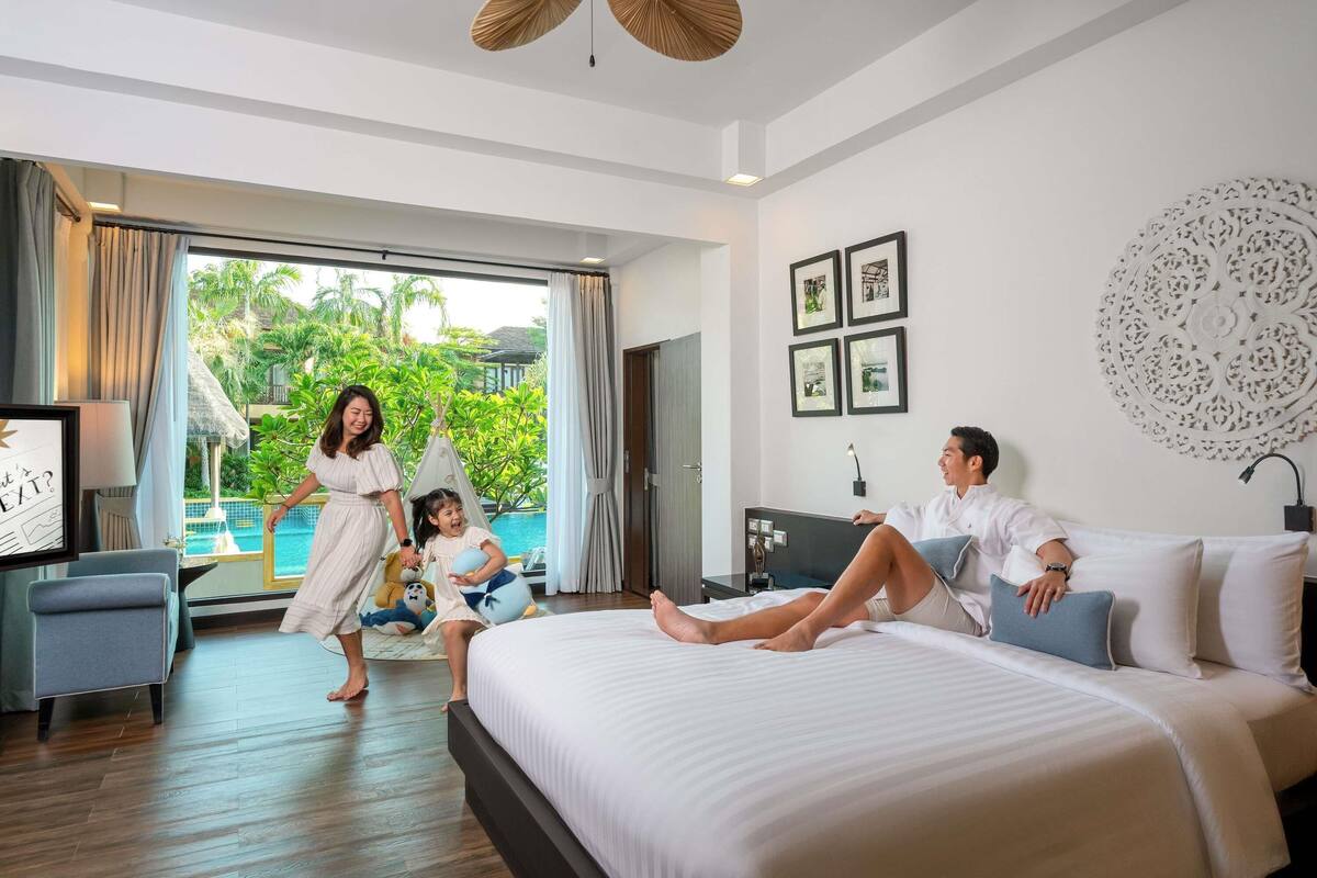 A hotel room with a bed, a desk, a chair, a window with a view of greenery, and a ceiling fan.