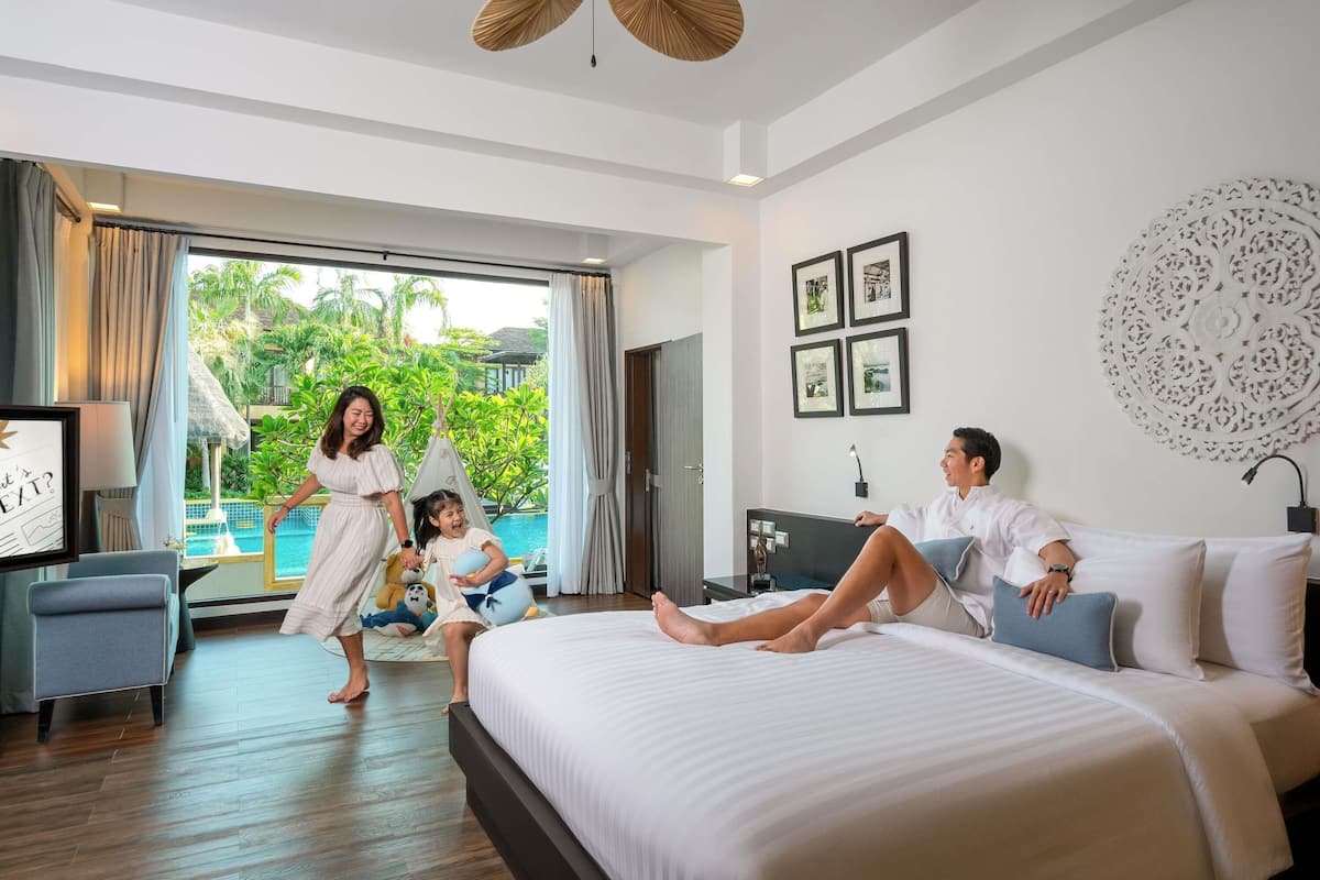 A hotel room with a bed, a desk, a chair, a window with a view of greenery, and a ceiling fan.