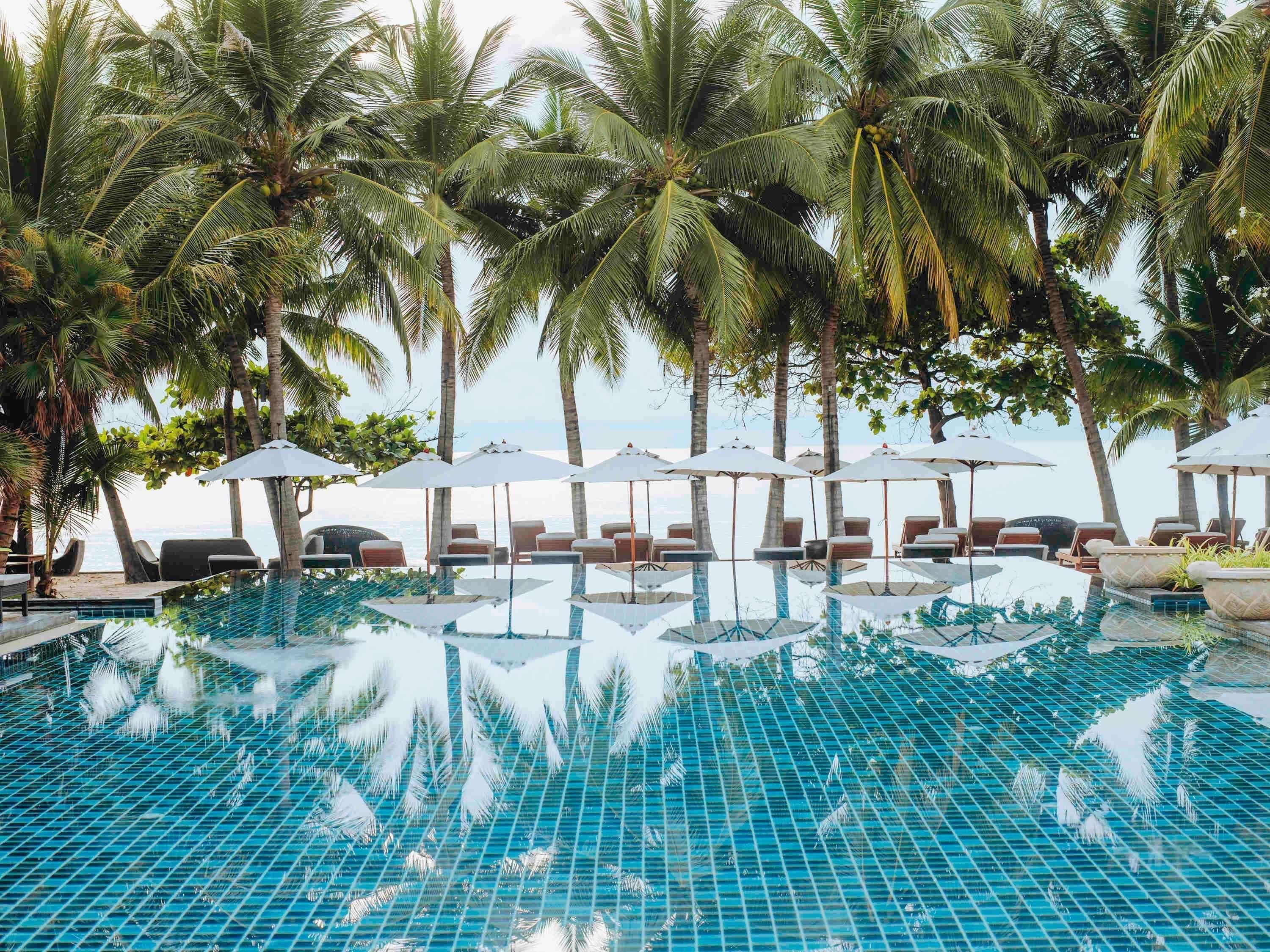 A pool with clear blue water, surrounded by lounge chairs and umbrellas, with palm trees and a view of the ocean.