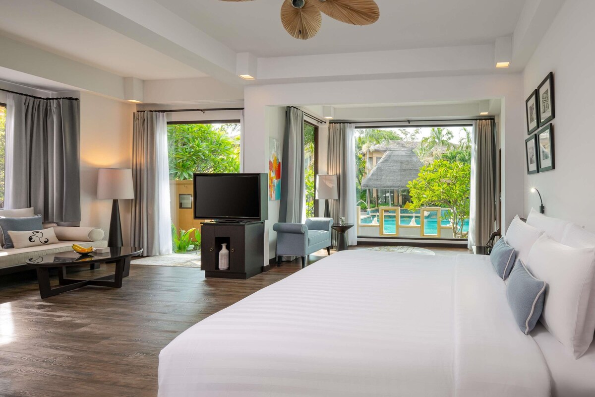 A spacious hotel room with a large bed, a flat-screen TV, a seating area with a sofa and armchairs, and a balcony with a pool view.