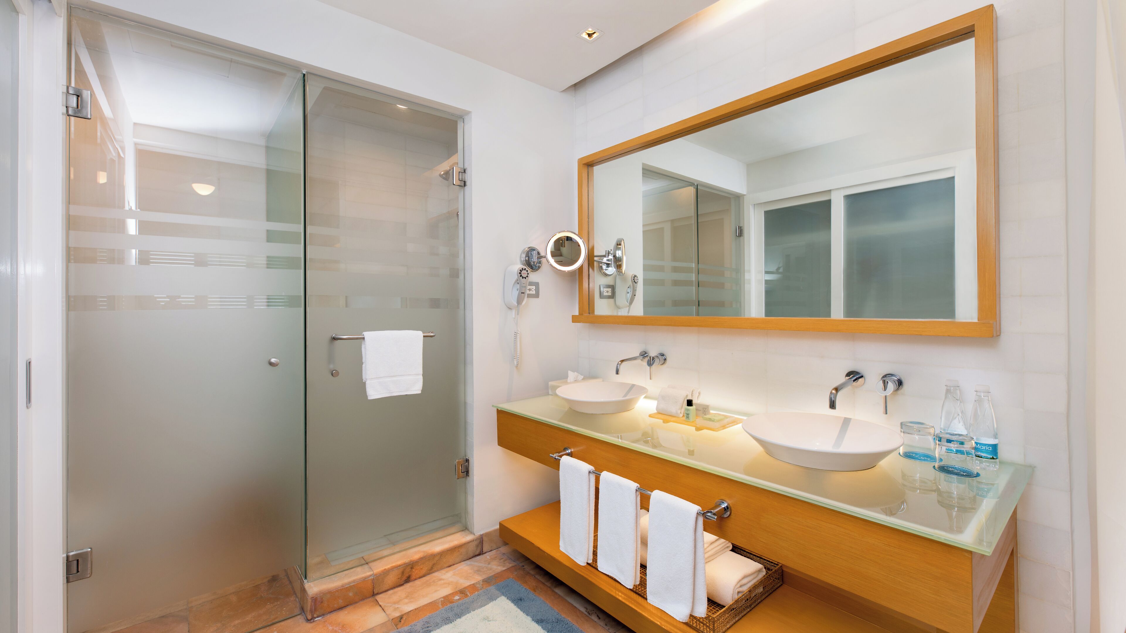 A modern bathroom with a glass shower door, a large mirror, two sinks, and a vanity with a marble countertop.