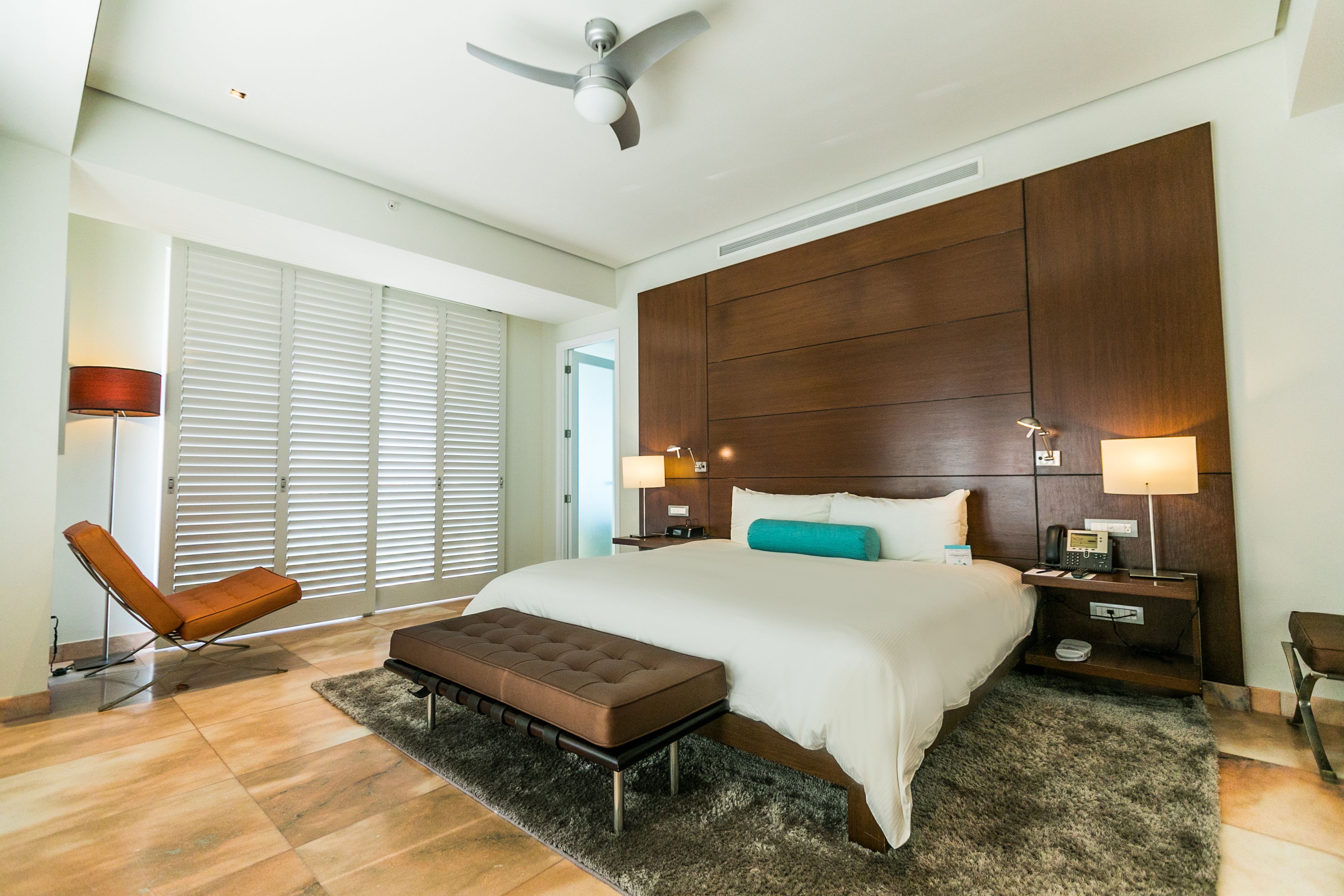 Een moderne hotelkamer met een groot bed, een houten hoofdbord, een plafondventilator en een zithoek met een fauteuil en voetenbankje.