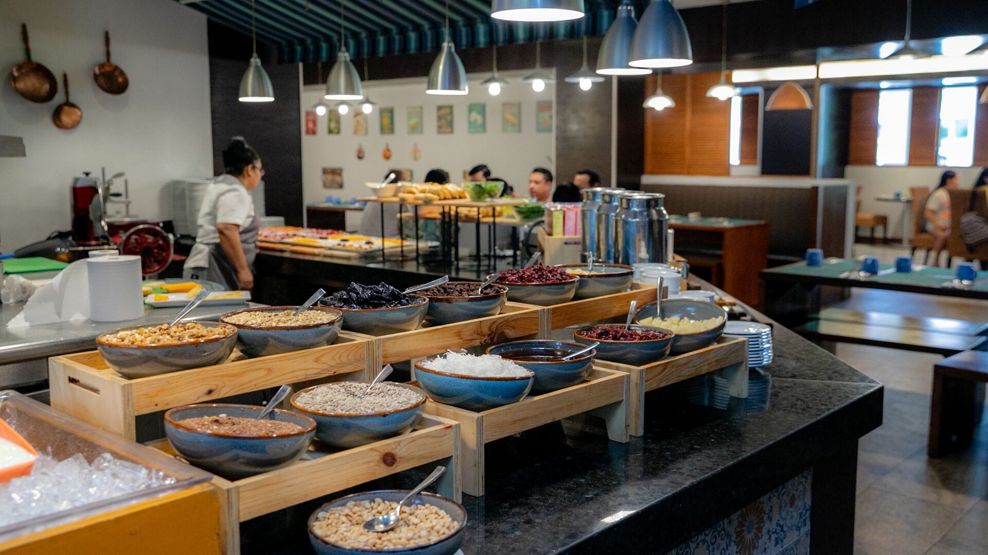 A buffet area with various food options in bowls and containers, a person serving food, and a counter with stacked plates.