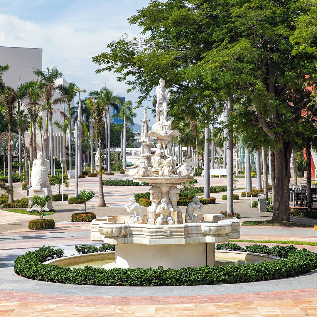 Una plaza pública con una fuente central, rodeada de palmeras y estatuas.